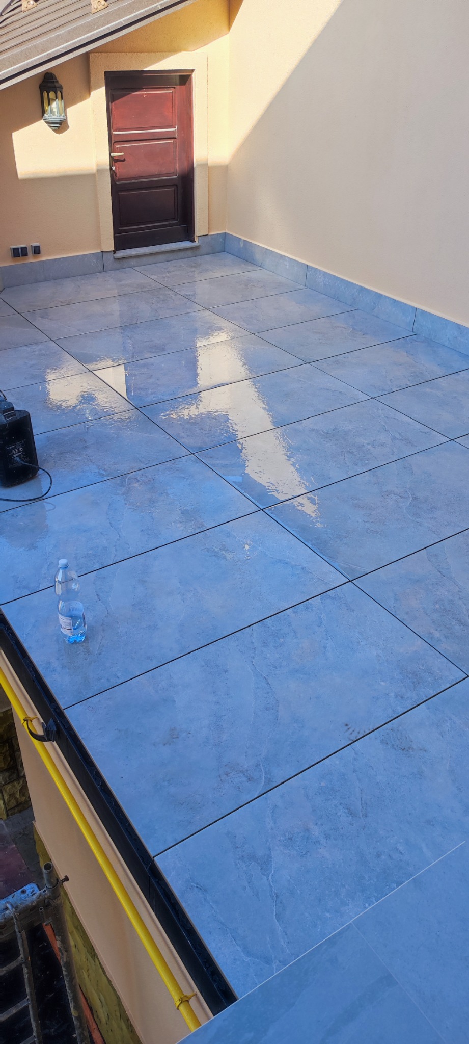 Wyłożony płytkami taras na poddaszu z widoczną butelką wody, lampą zewnętrzną i drzwiami wejściowymi.