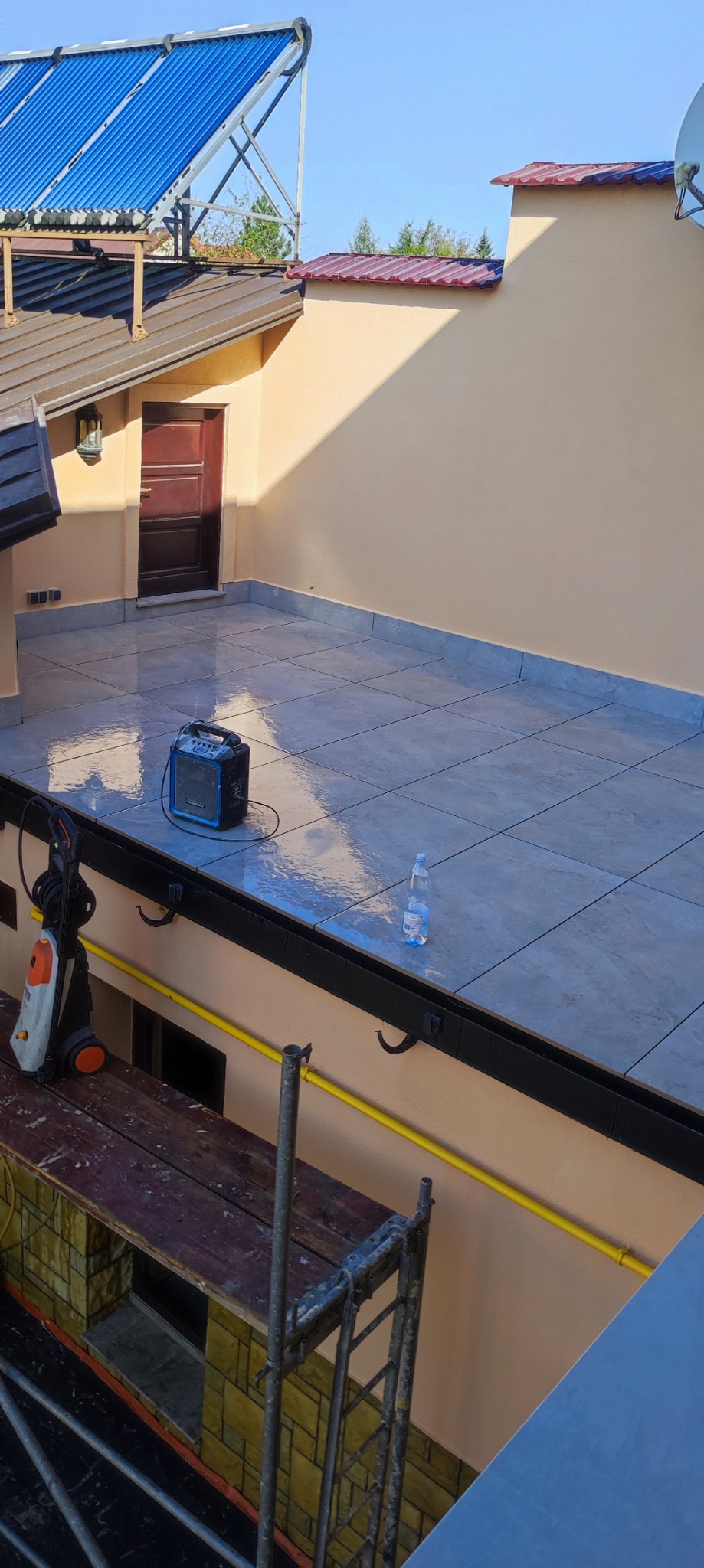 Wykafelkowany taras na dachu budynku, z widocznymi śladami wilgoci po czyszczeniu, przenośny głośnik Bluetooth, butelka wody, myjka ciśnieniowa i rusztowanie.