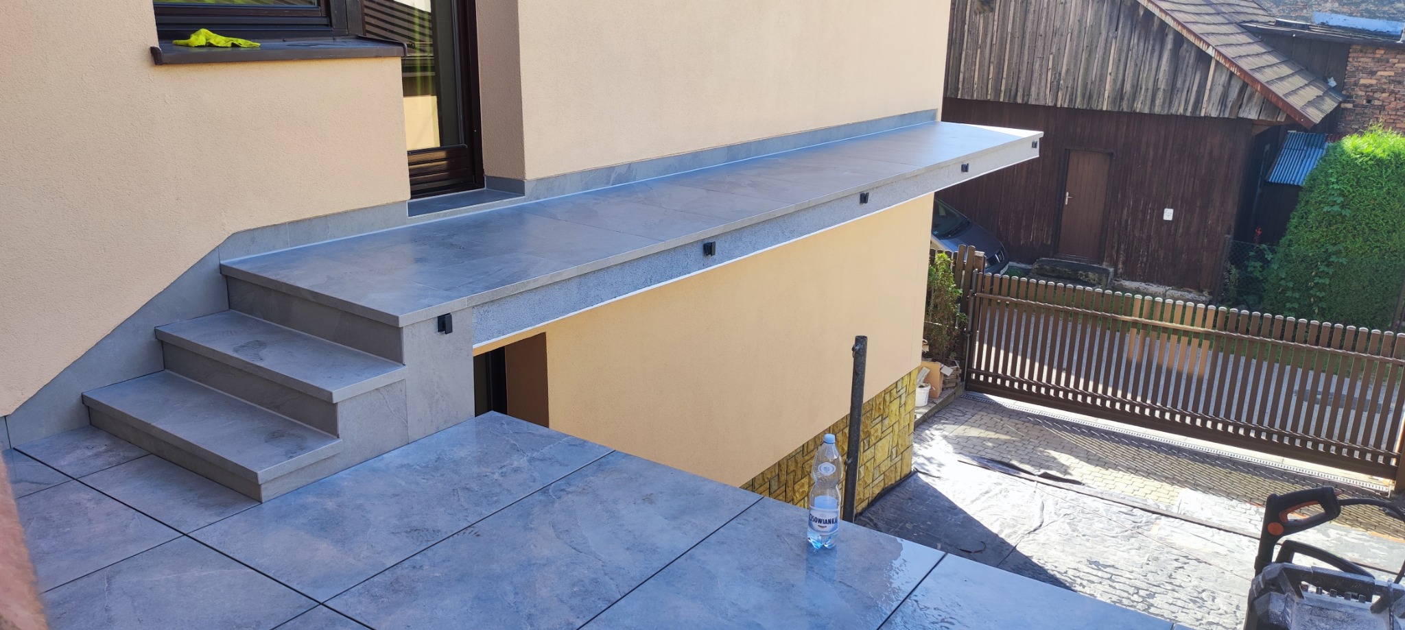 Wykończony taras z szarych płytek z widocznymi schodami, zadaszeniem i elewacją budynku w kolorze beżowym, butelka wody mineralnej na płytkach tarasu.