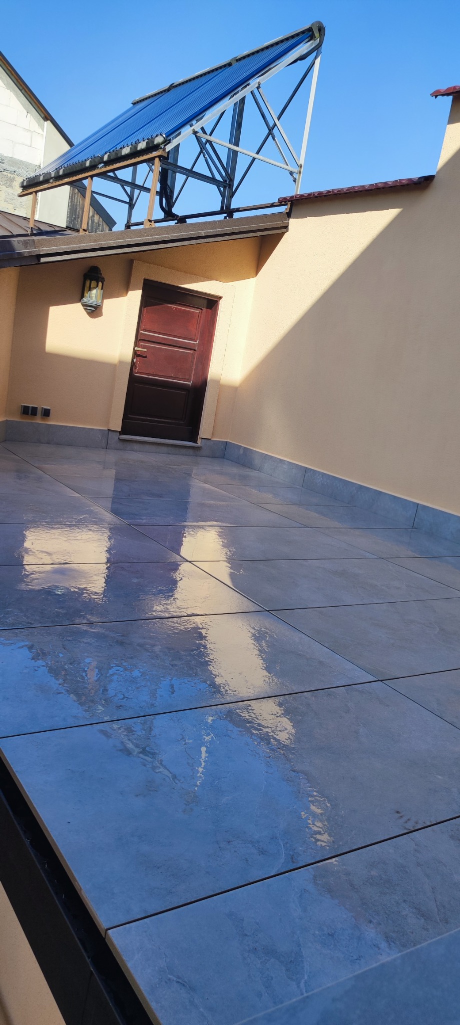 Błyszcząca, szara posadzka z płytek ceramicznych na tarasie, z widocznym odbiciem ściany i drzwi wejściowych oraz instalacji solarnych na dachu.