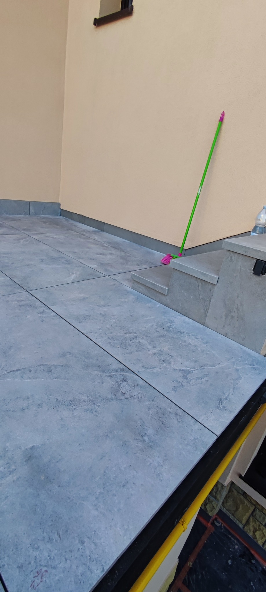 Wyłożona szarymi płytkami podłoga na zewnątrz budynku, widoczna zielona miotła oparta o beżową ścianę i butelka wody na schodku.