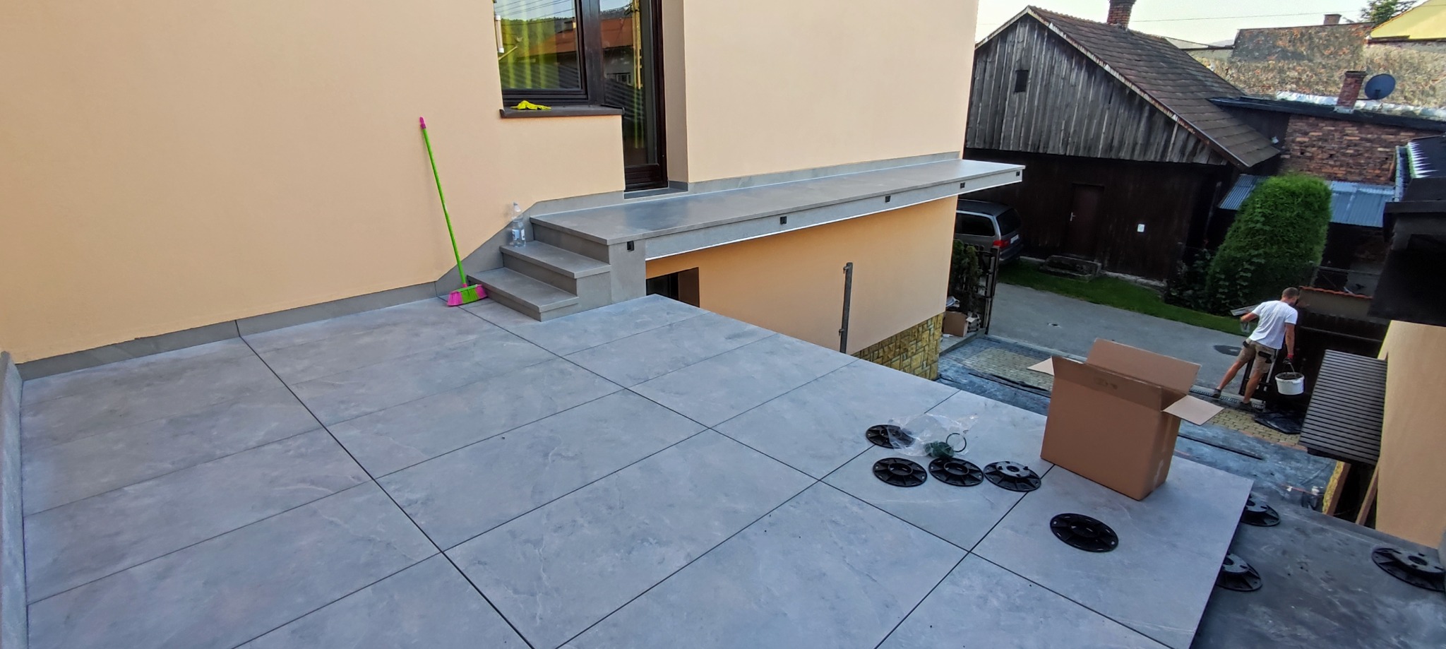 Montaż płytek na tarasie na regulowanych wspornikach, widoczne elementy montażowe, karton i pracownik w tle.