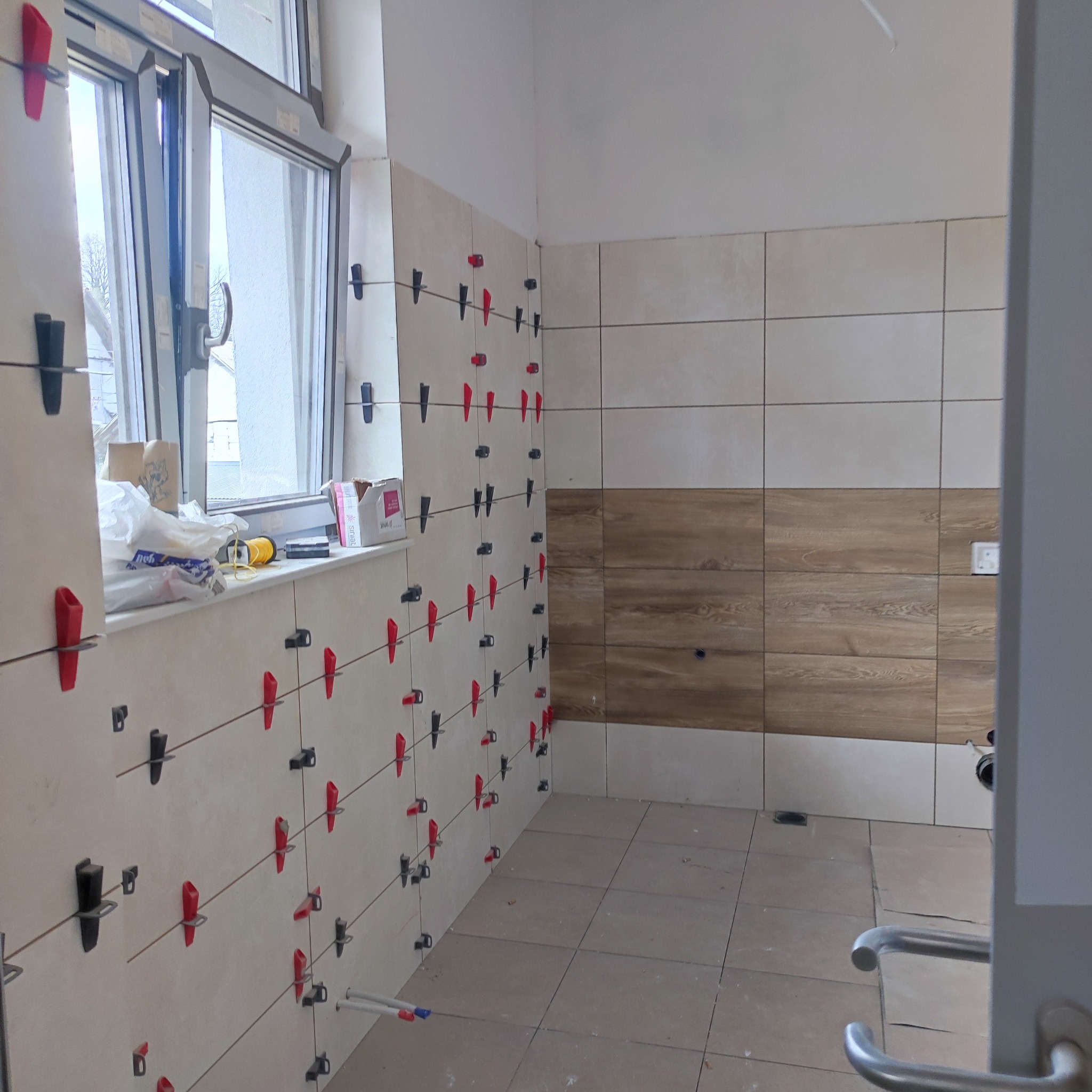 Ściana i podłoga w trakcie układania płytek ceramicznych, z użyciem czerwono-czarnych klipsów poziomujących, widoczne narzędzia na parapecie okiennym.