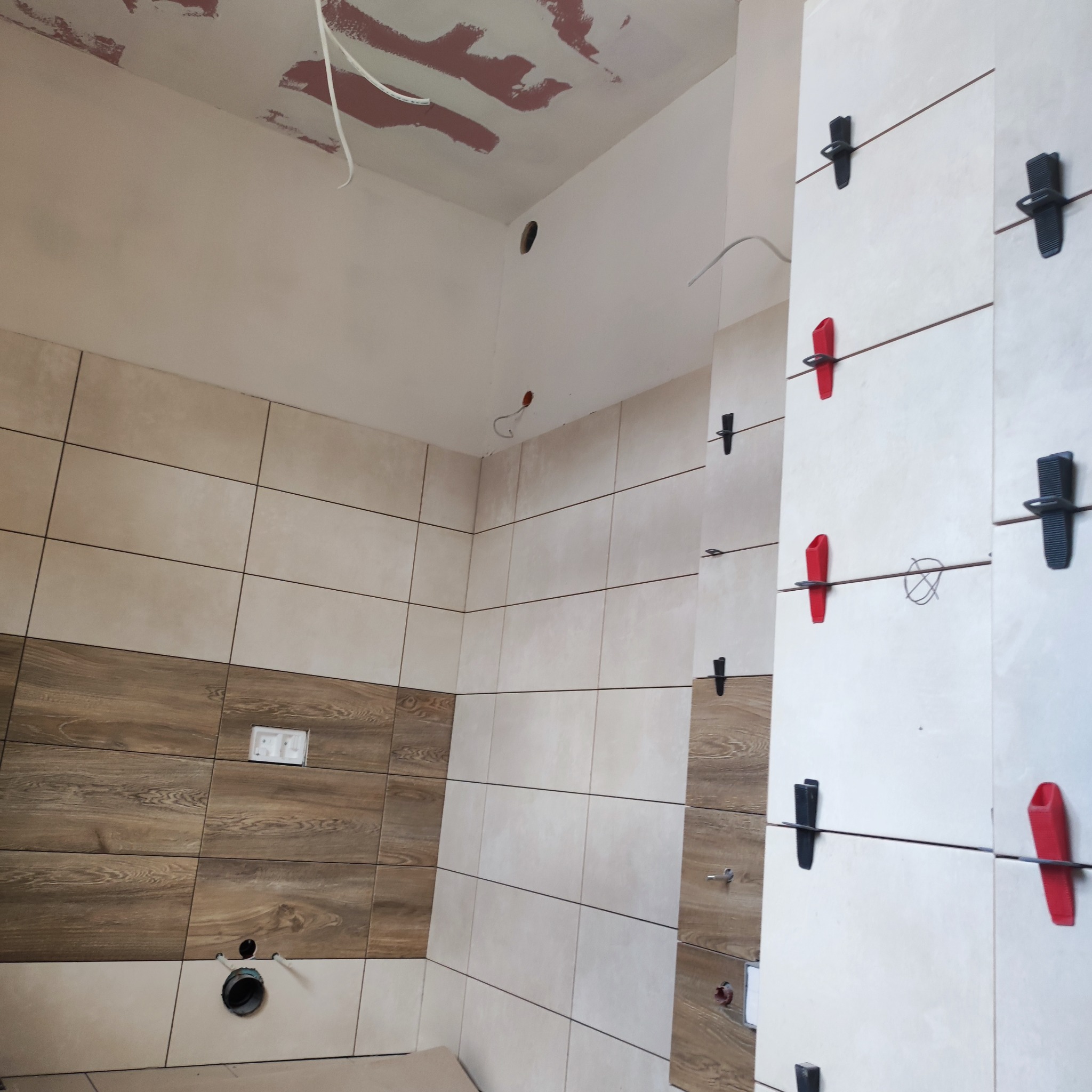 Ściana w trakcie układania płytek z użyciem klipsów poziomujących, częściowo wykończona, z widocznymi otworami na instalacje i niedomalowanym sufitem.