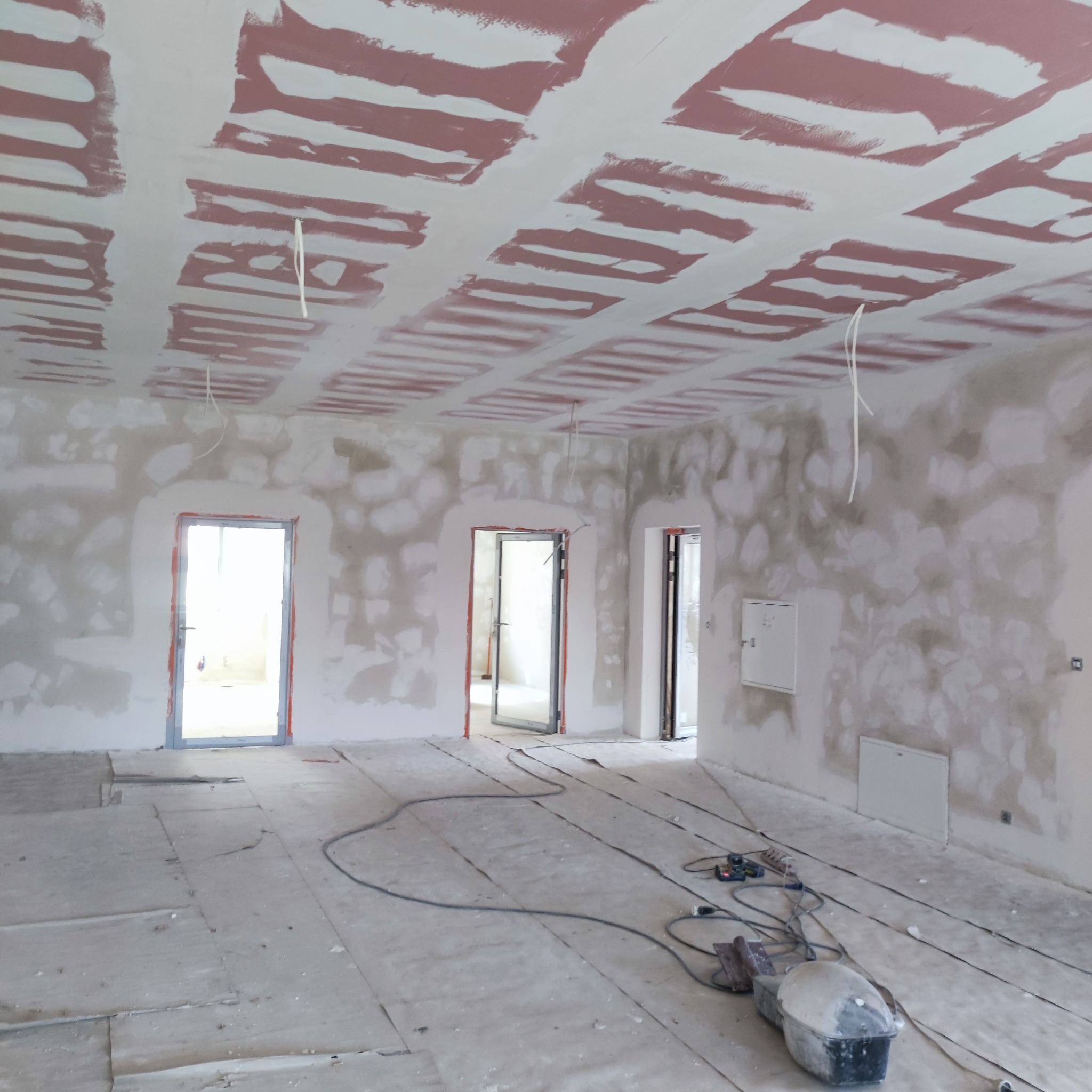 Wnętrze w trakcie prac wykończeniowych: sufit z płyt kartonowo-gipsowych z widocznymi łączeniami pokrytymi szpachlą, ściany częściowo przygotowane pod malowanie, widoczne kable i narzędzia...