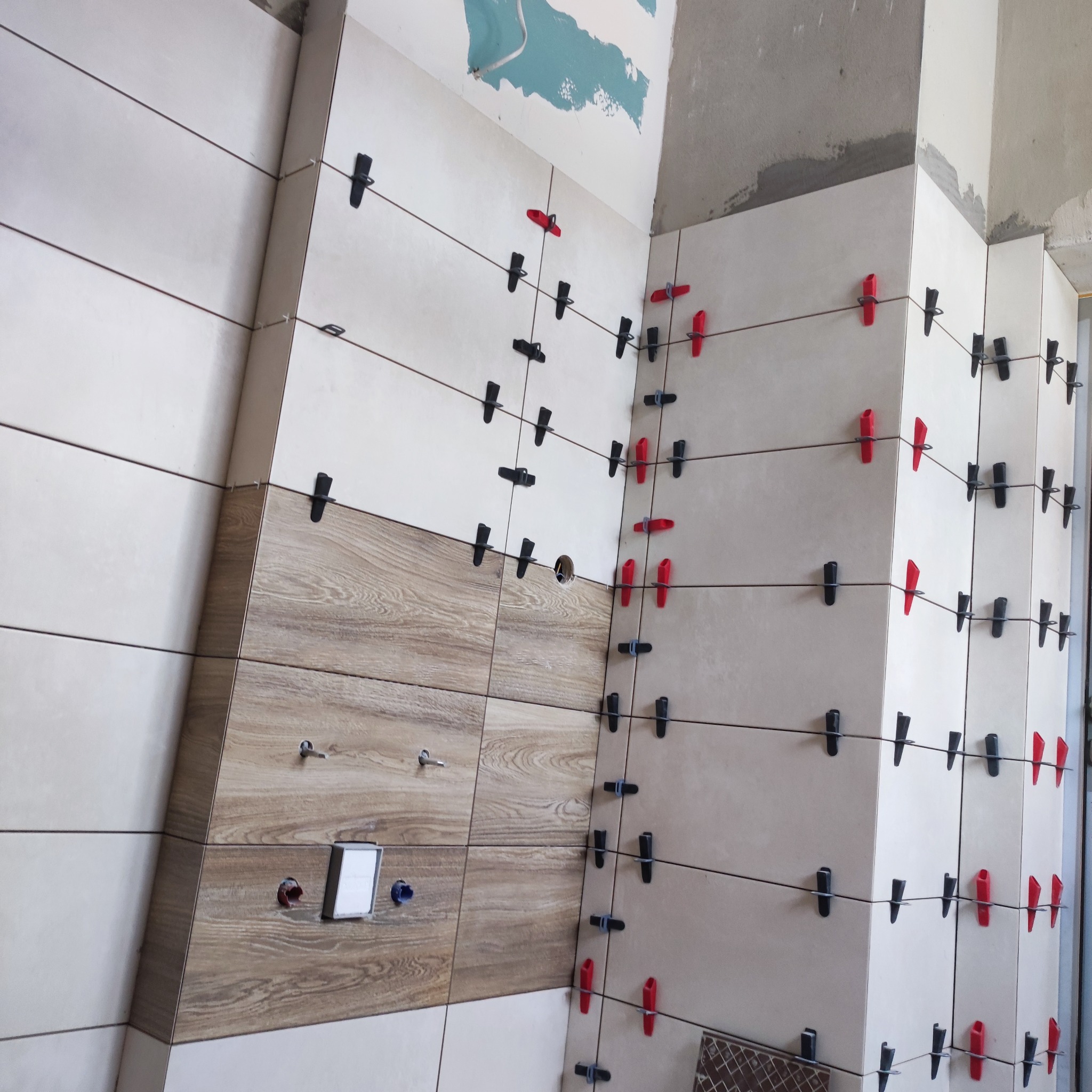 Układanie płytek ściennych z użyciem klipsów poziomujących, widoczne fragmenty ściany z instalacją hydrauliczną i elektryczną.