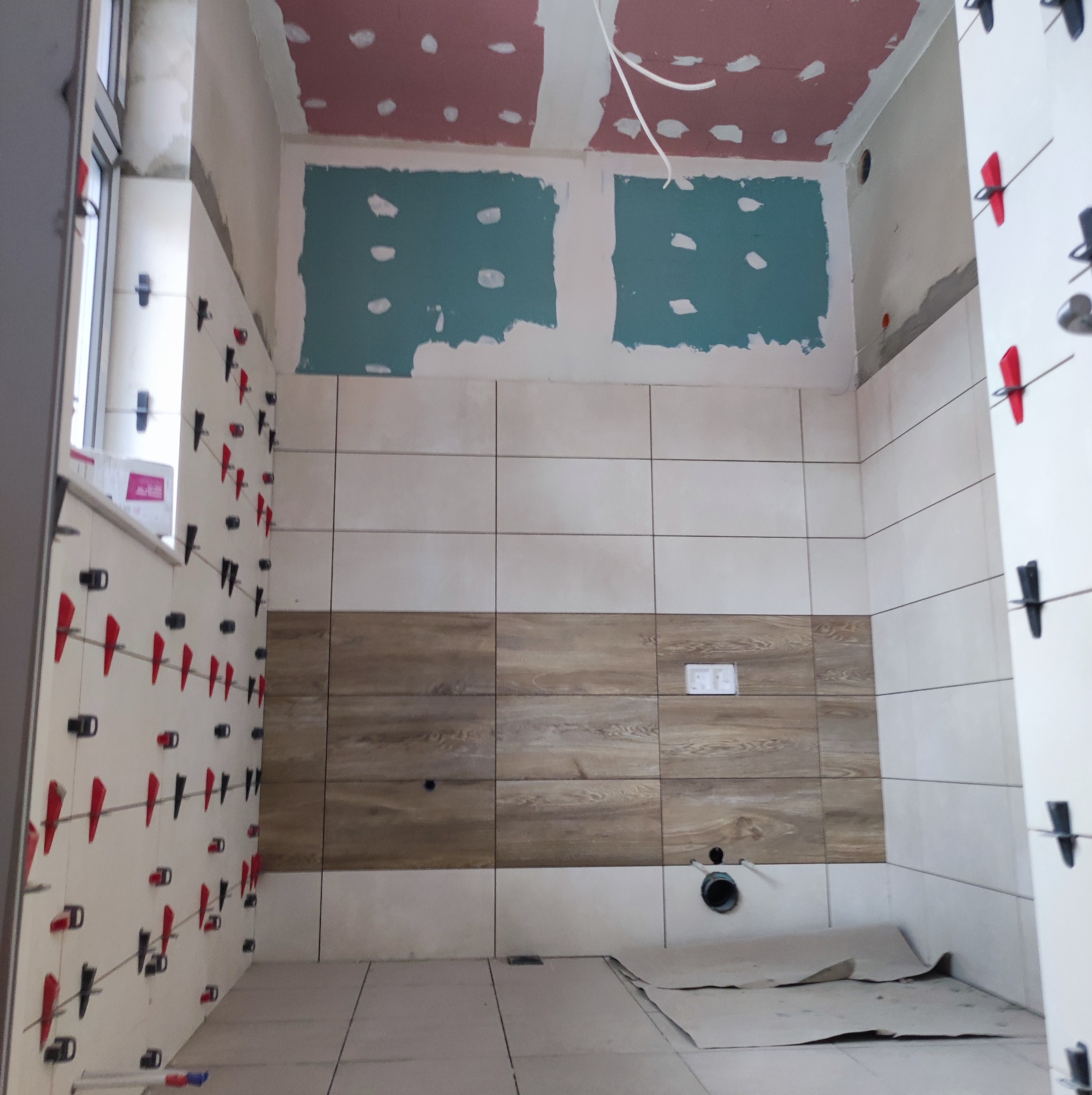 Wnętrze łazienki w trakcie prac wykończeniowych: ułożone płytki ścienne z poziomym pasem płytek imitujących drewno, ściany częściowo pokryte płytami kartonowo-gipsowymi, na ścianach widoczne klipsy...