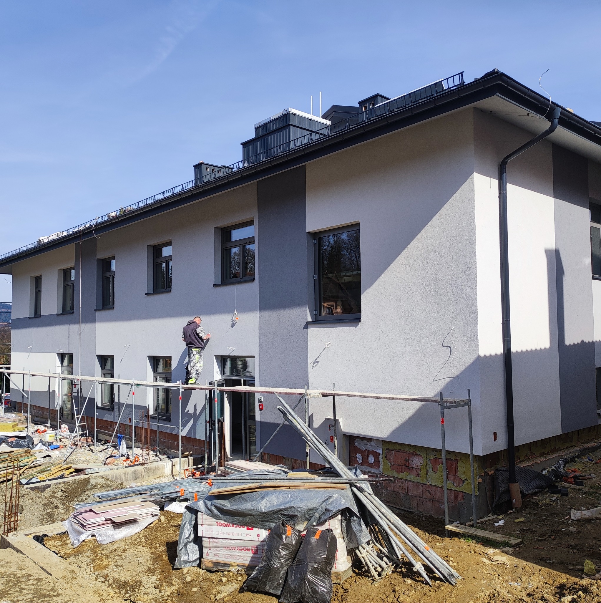 Pracownik na rusztowaniu maluje elewację dwukondygnacyjnego budynku na szaro, widoczne materiały budowlane i rusztowania.