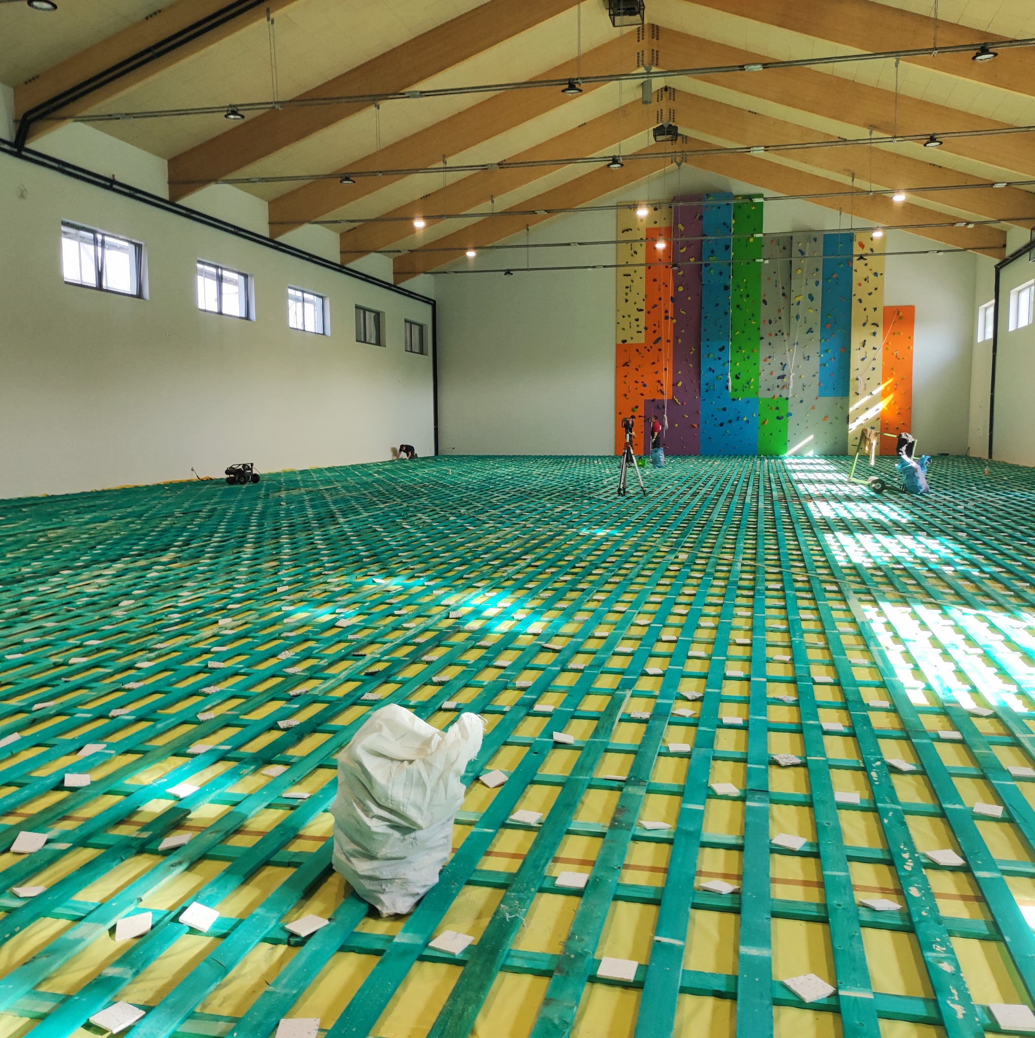 Przygotowanie podłoża pod panele lub płytki w dużej hali sportowej: zielona kratownica z drewnianych belek na żółtej folii, białe płytki dystansowe, w tle ściana wspinaczkowa i pracownicy.