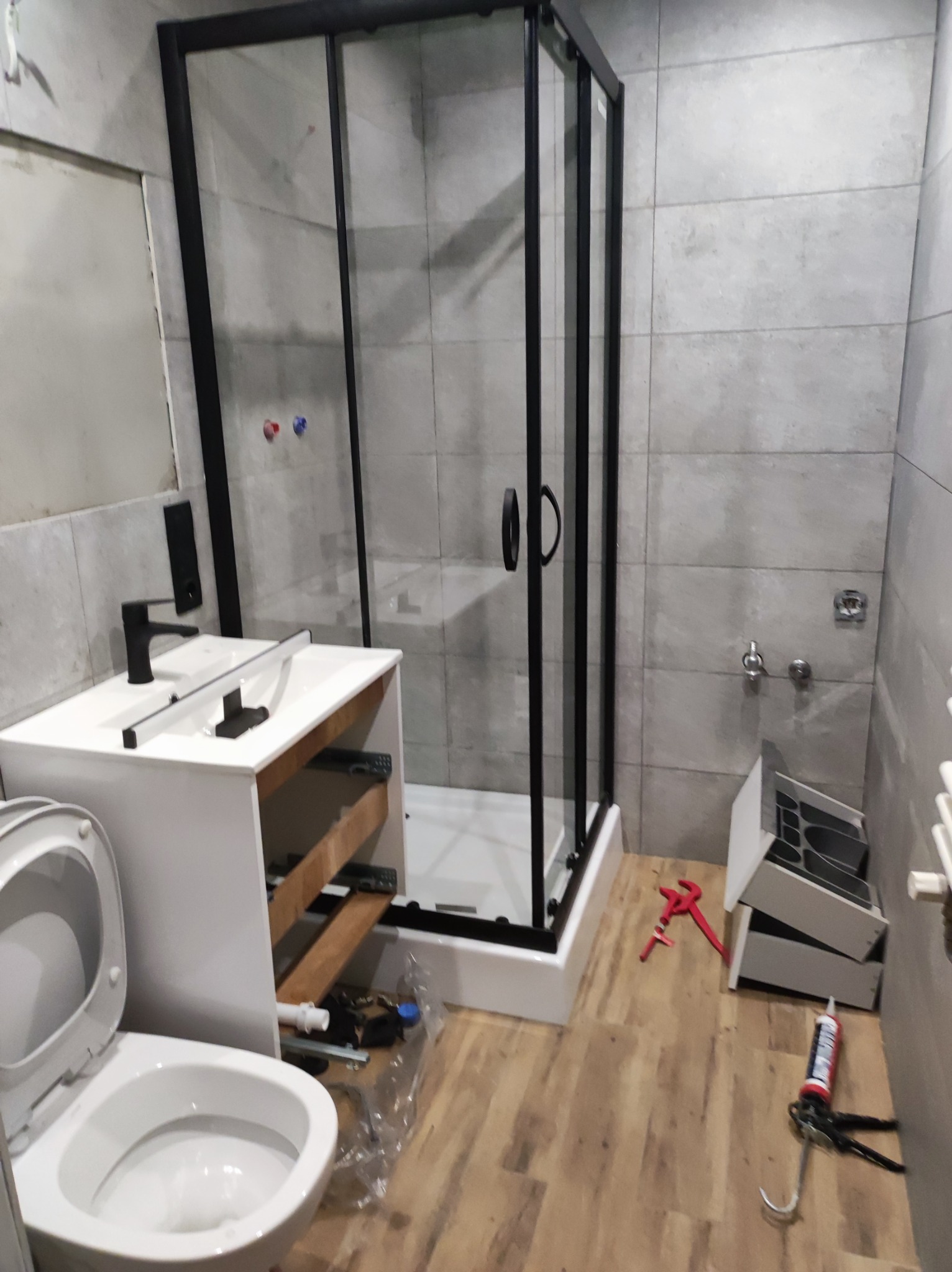 Montaż kabiny prysznicowej z czarnymi ramami i białym brodzikiem w łazience z szarymi płytkami, widoczna toaleta i niedokończona szafka pod umywalkę z wyciągniętymi szufladami, narzędzia i silikon...