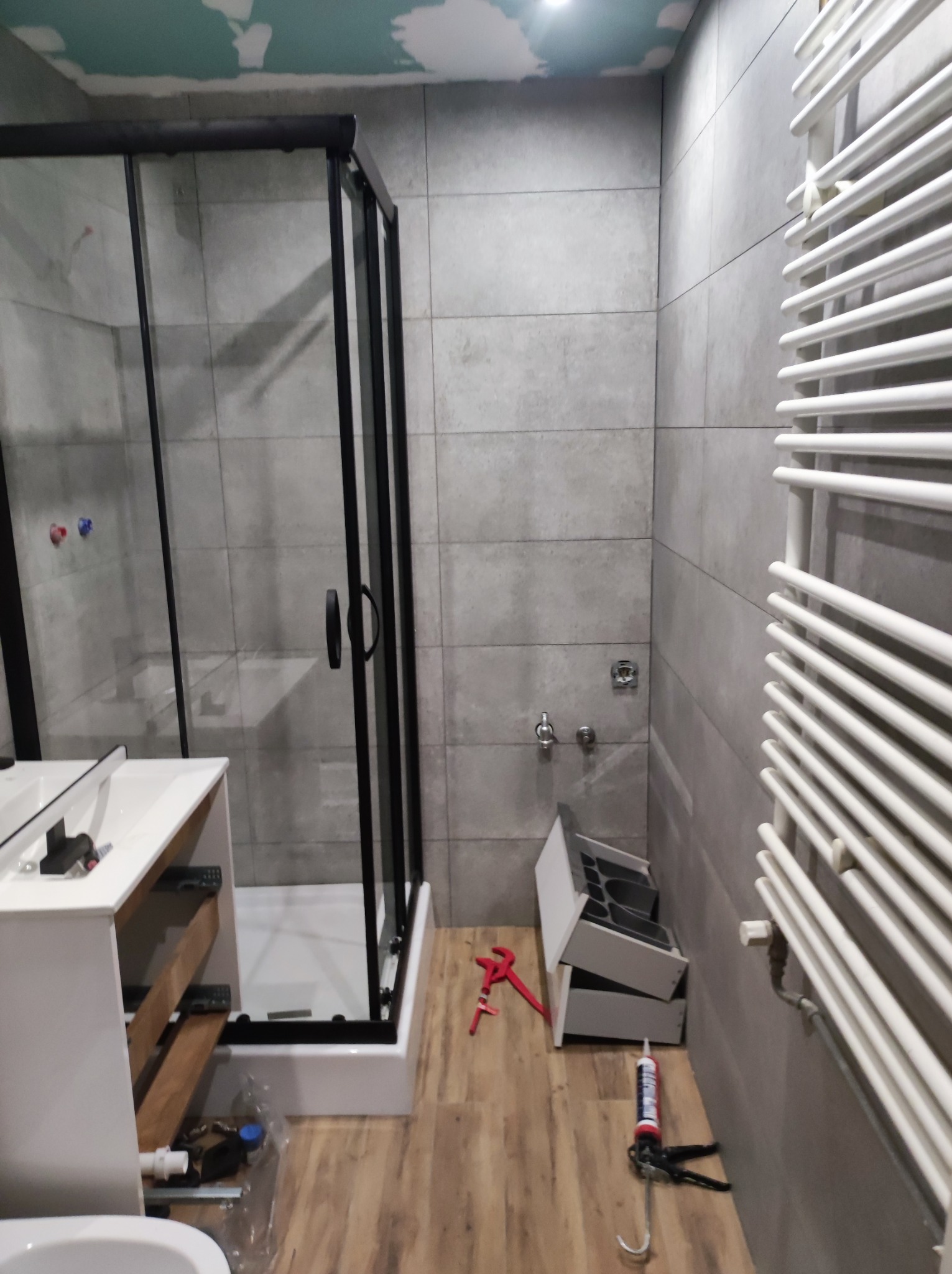 Łazienka w trakcie prac wykończeniowych: kabina prysznicowa z czarnymi profilami, biała szafka pod umywalkę z otwartymi szufladami, grzejnik drabinkowy, szare płytki na ścianach, widoczne narzędzia...