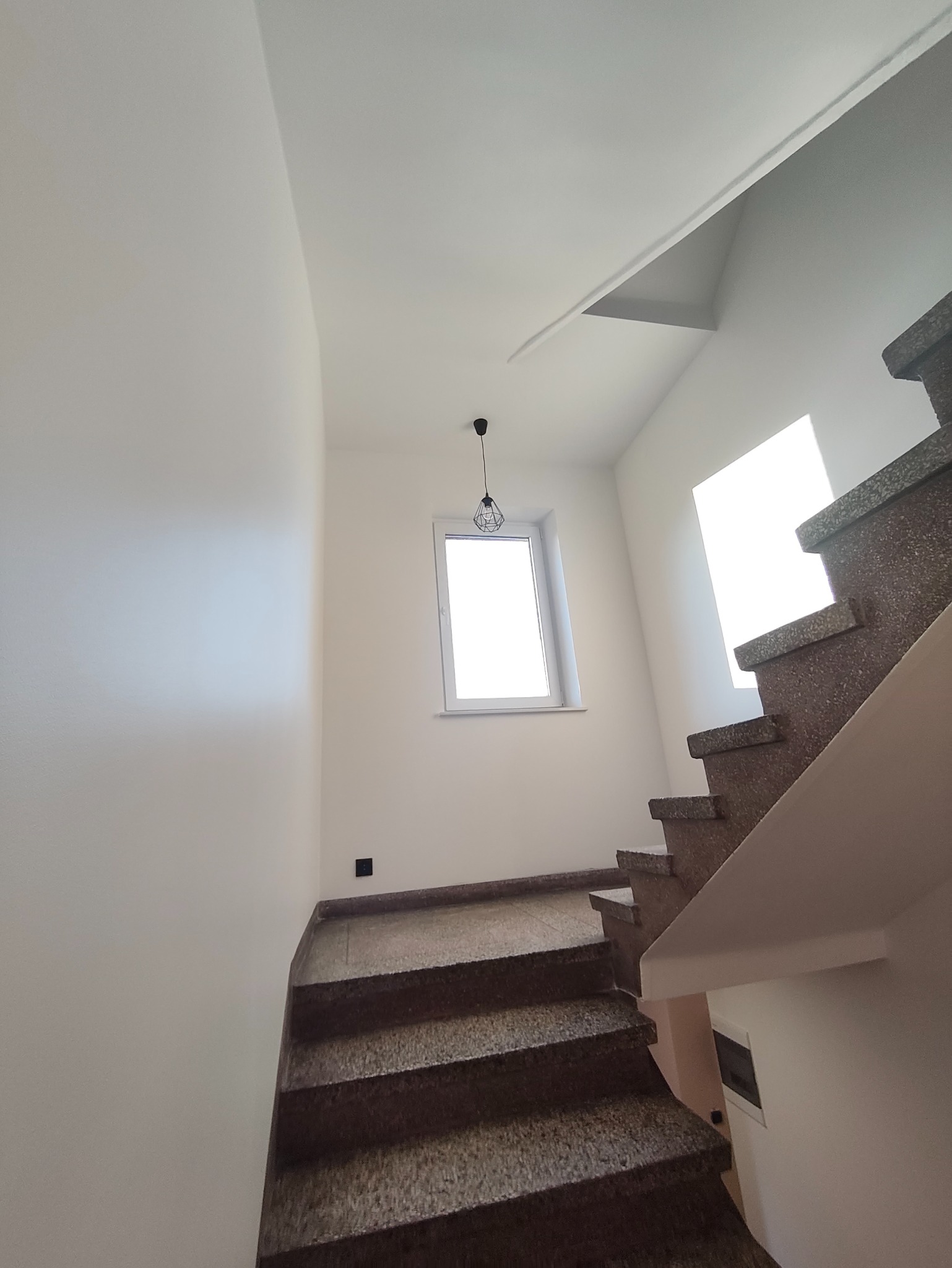 Klatka schodowa po remoncie: świeżo pomalowane ściany w odcieniach bieli, granitowe schody i nowoczesna lampa wisząca. Widok z dołu, podkreślający przestrzeń i detale wykończenia.
