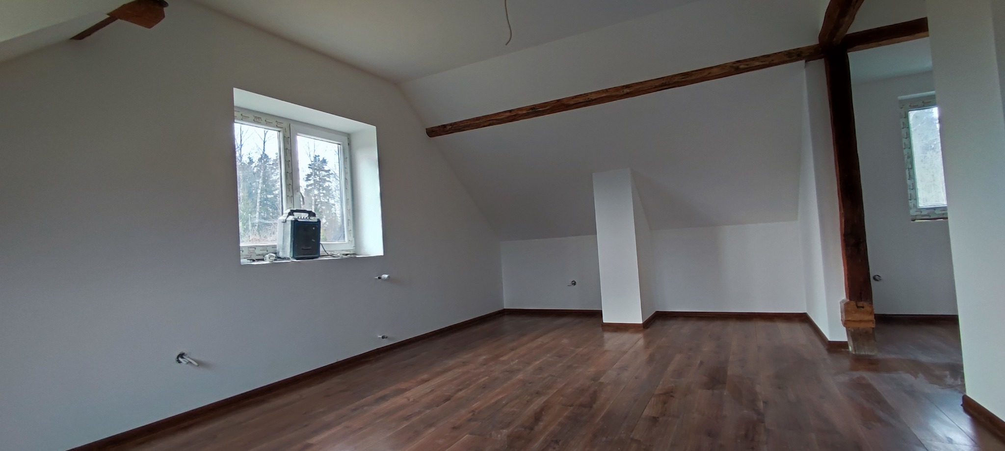 Poddasze po remoncie: brązowe panele, białe ściany, drewniane belki stropowe. Widoczne gniazdka elektryczne bez osprzętu i okno z widokiem na drzewa.