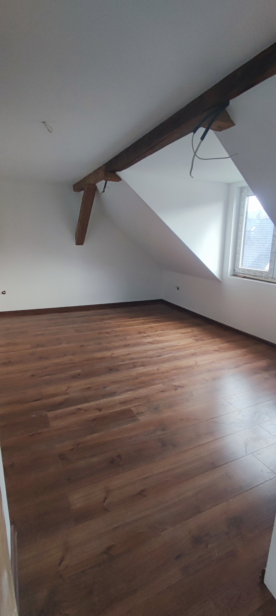 Poddasze po remoncie: brązowe panele podłogowe, białe ściany i skośny sufit z drewnianą belką. Widoczne okno. Nowoczesne wykończenie wnętrza.
