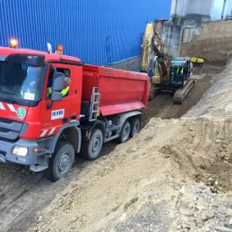 Czerwona wywrotka Mercedes-Benz HAWI z kierowcą w kamizelce odblaskowej i włączonym pomarańczowym światłem ostrzegawczym, na nierównym terenie z ziemią i kamieniami, w tle koparka i niebieska hala.