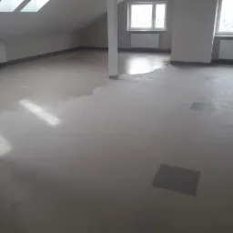 Wyrównana, szara wylewka samopoziomująca w jasnym, przestronnym pomieszczeniu na poddaszu z oknami dachowymi i grzejnikami, widoczne kwadratowe ślady po zabezpieczeniach.