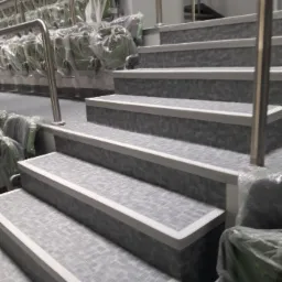 Schody wykończone szarą, wzorzystą okładziną z białymi krawędziami, prowadzące do rzędu krzeseł przykrytych folią ochronną, widoczne metalowe poręcze.