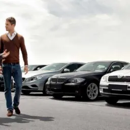 Dwóch mężczyzn w garniturze i swetrze rozmawia na tle rzędu zaparkowanych samochodów różnych marek na betonowym placu w pochmurny dzień.