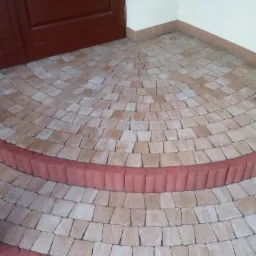 Półokrągłe schody wejściowe wyłożone beżową kostką brukową i obrzeżami z czerwonych elementów betonowych, prowadzące do ciemnych, drewnianych drzwi.