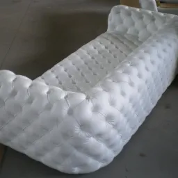 Biała sofa Chesterfield z głębokimi pikowaniami na gładkiej, szarej posadzce, widoczna z boku, w tle materiały tapicerskie.