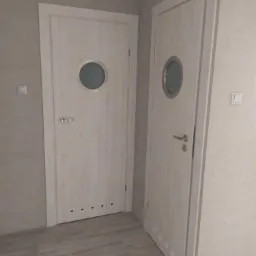 Dwie białe, drewnopodobne drzwi wewnętrzne z okrągłymi przeszkleniami w stylu bulajów, zamontowane w szarej ścianie i z szarą podłogą.