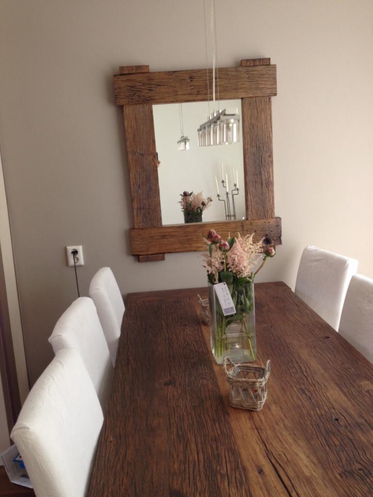 Stół z litego drewna z białymi krzesłami, nad nim lustro w drewnianej ramie, odbijające wiszącą lampę i świeczniki, na stole wazon z kwiatami.