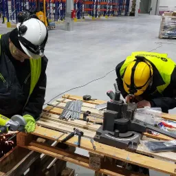 Dwóch monterów w kaskach i kamizelkach odblaskowych pracuje nad metalowymi elementami w hali magazynowej, jeden szlifuje kątówką z iskrami, drugi używa giętarki do metalu, na palecie narzędzia...