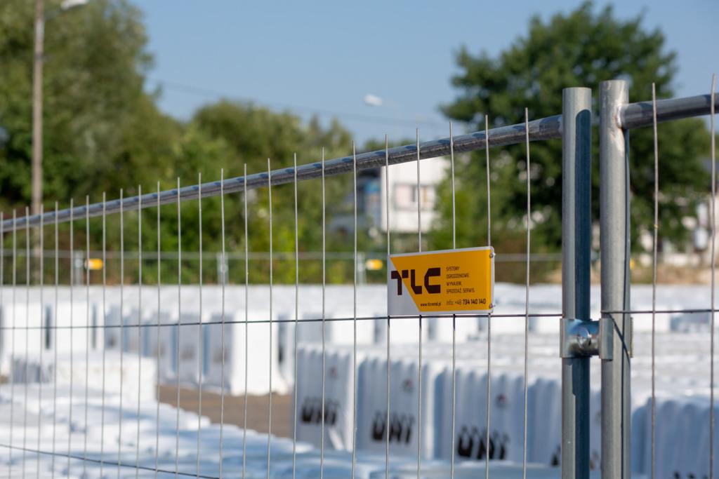 Tymczasowe ogrodzenie budowlane z logo firmy TLC na żółtej tabliczce, widoczne białe betonowe podstawy ogrodzenia w tle.