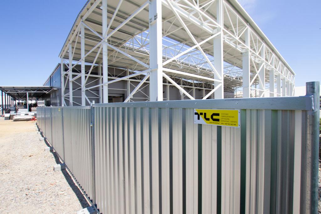 Tymczasowe ogrodzenie budowlane z blachy falistej, z żółtą tabliczką informacyjną TLC, na tle konstrukcji stalowej hali w budowie.
