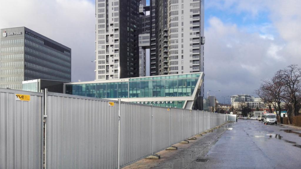Tymczasowe ocynkowane ogrodzenie panelowe z widocznymi budynkami w tle, w tym wieżowiec Sea Towers w Gdyni, na mokrej nawierzchni po deszczu.