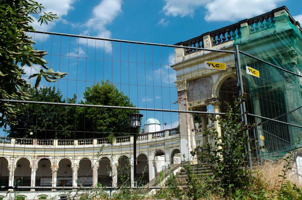 Zabezpieczony siatką plac budowy przed zabytkowym budynkiem z kolumnami i arkadami, widoczne logo TLC na siatce.