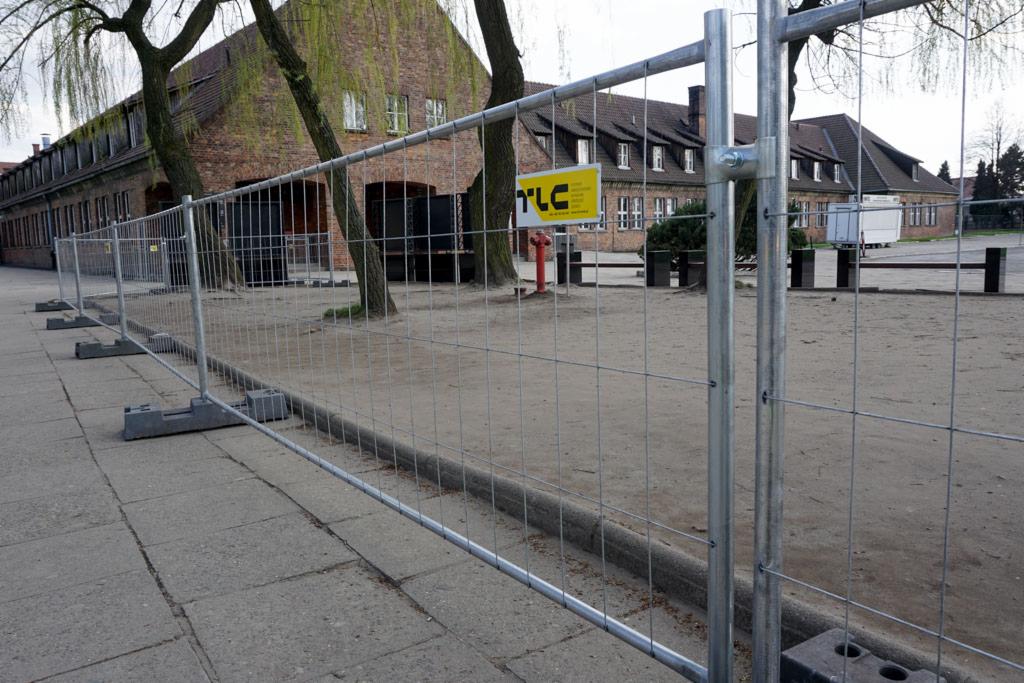 Tymczasowe ogrodzenie z siatki na betonowych podstawach, widoczne budynki z czerwonej cegły w tle oraz hydrant przeciwpożarowy z żółtą tabliczką.