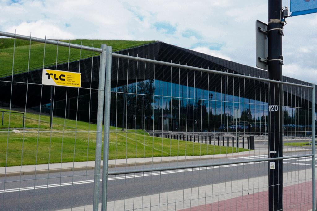 Tymczasowe ogrodzenie budowlane z żółtą tabliczką informacyjną na tle nowoczesnego budynku z zielonym dachem.