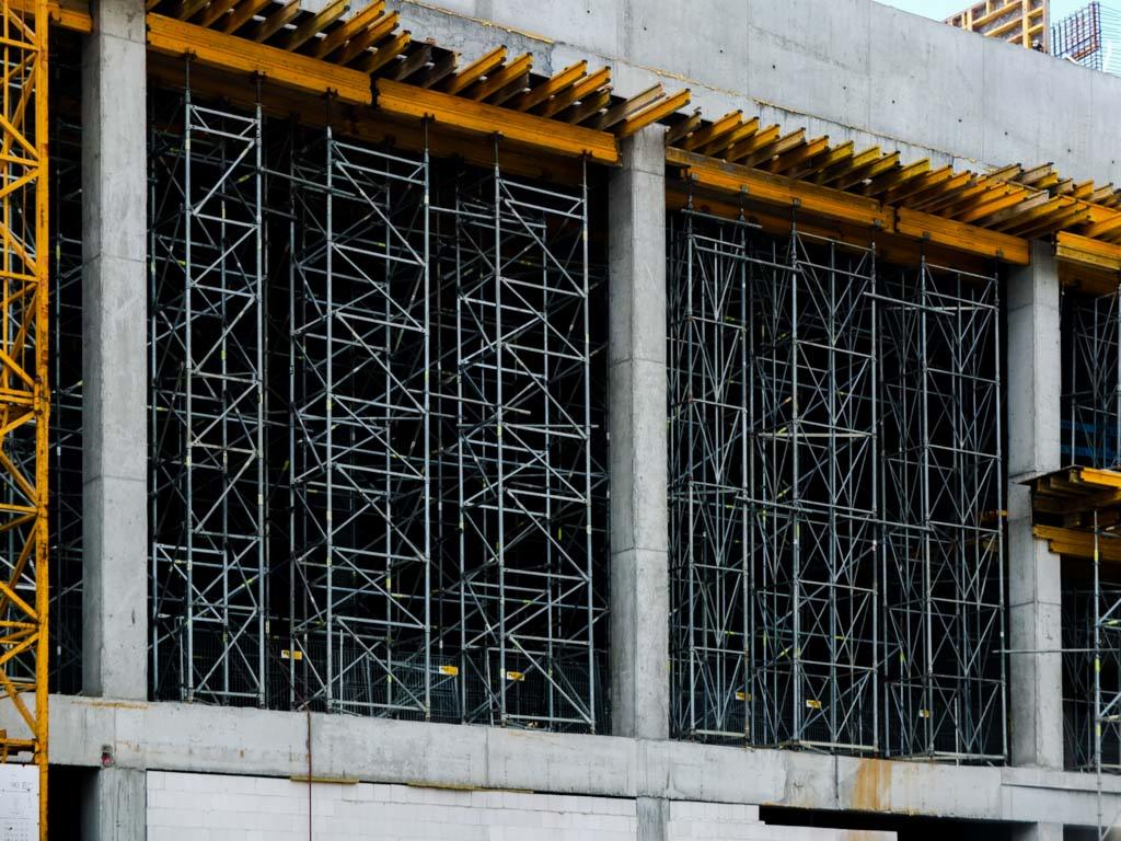 Szare betonowe ściany budynku w trakcie budowy, z widocznymi metalowymi rusztowaniami i żółtym dźwigiem po lewej stronie.