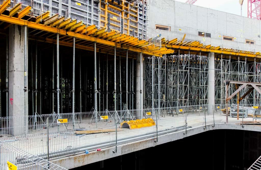 Szare betonowe filary podtrzymujące żółte belki stropowe na placu budowy, widoczne rusztowania i elementy konstrukcyjne w tle.