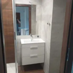 Nowoczesna łazienka z szarymi płytkami, drewnianą podłogą i białą szafką pod umywalką; widoczne przewody wystające ze ściany nad lustrem sugerują niedokończony montaż oświetlenia.