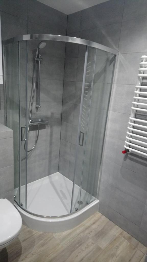 Narożna kabina prysznicowa z chromowanymi profilami i białym brodzikiem, zamontowana w łazience w Ostrołęce wykończonej szarymi płytkami imitującymi kamień i panelami podłogowymi w odcieniach drewna.