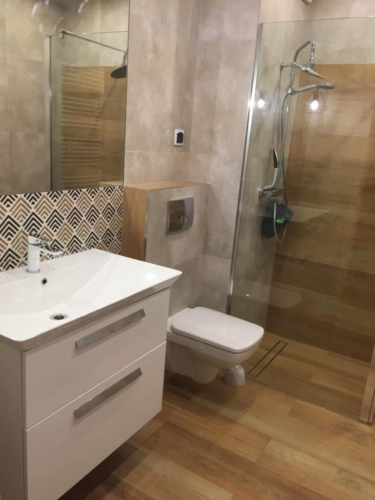 Nowoczesna łazienka z drewnopodobną podłogą, białą szafką pod umywalką z geometrycznym wzorem na blacie, wiszącą toaletą i kabiną prysznicową ze szklaną ścianką.