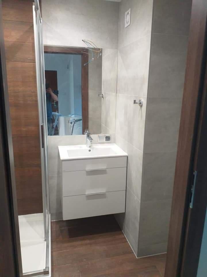 Nowoczesna łazienka z szarymi płytkami, drewnianą podłogą i białą szafką pod umywalką; widoczne przewody wystające ze ściany nad lustrem sugerują niedokończony montaż oświetlenia.