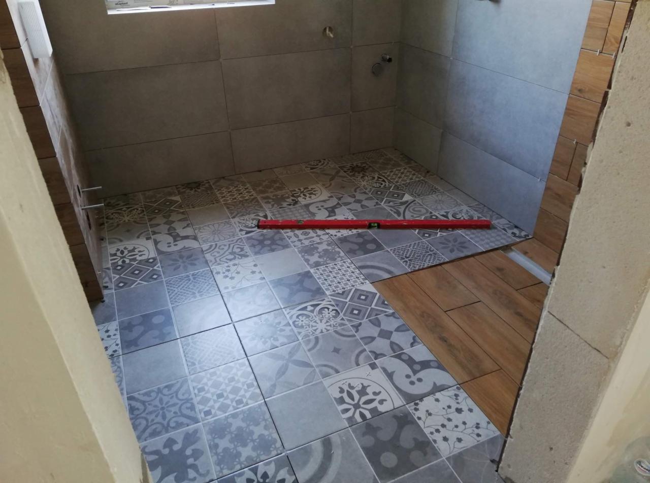 Układanie płytek podłogowych w łazience, widoczny poziomica na szarych płytkach wzorzystych i częściowo ułożone płytki imitujące drewno.