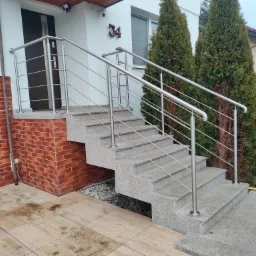 Zewnętrzne schody z szarego kamienia z prostą, metalową barierką ze stali nierdzewnej, prowadzące do drzwi wejściowych domu z numerem 34, otoczone tujami.