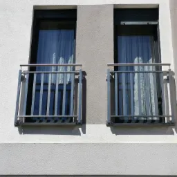 Dwa balkony z prostymi, metalowymi barierkami na tle białej elewacji budynku z widocznymi zasłonami w oknach.