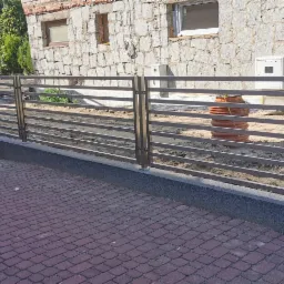 Nowoczesne ogrodzenie z poziomych metalowych profili, widok z zewnątrz posesji, na pierwszym planie czerwona kostka brukowa.