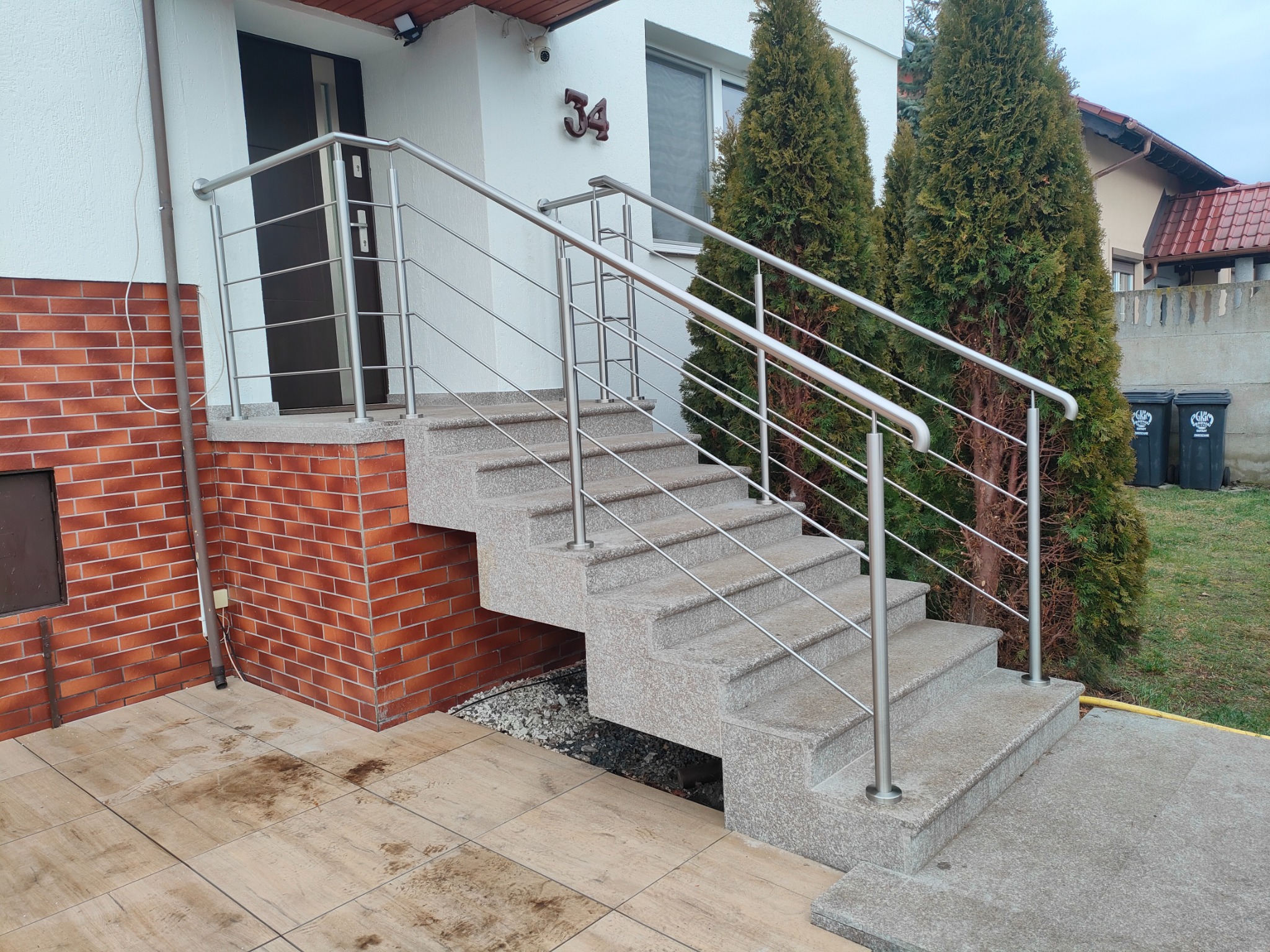 Zewnętrzne schody z szarego kamienia z prostą, metalową barierką ze stali nierdzewnej, prowadzące do drzwi wejściowych domu z numerem 34, otoczone tujami.