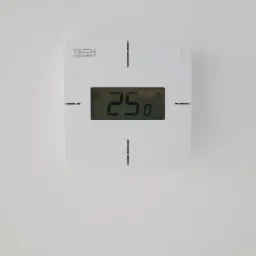 Biały, kwadratowy termostat TECH na ścianie, wyświetlający temperaturę 25 stopni Celsjusza.