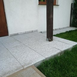 KTM-BUD JACEK LUBACZEWSKI - Wyłożony jasnoszarą, nakrapianą płytką taras z drewnianym słupem podtrzymującym zadaszenie, obok ściana budynku z oknem i trawnik.