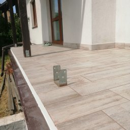 KTM-BUD JACEK LUBACZEWSKI - Wyłożony płytkami taras z widocznymi metalowymi wspornikami, świeżo wykończone obrzeża, drewnopodobne płytki w odcieniach beżu i brązu, fragment elewacji budynku w tle.