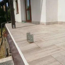 Wyłożony płytkami taras z widocznymi metalowymi wspornikami, świeżo wykończone obrzeża, drewnopodobne płytki w odcieniach beżu i brązu, fragment elewacji budynku w tle.