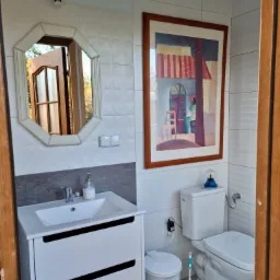 Jasna łazienka z białymi płytkami, umywalką z szafką w kolorze białym, toaletą i ozdobnym obrazem w drewnianej ramie na ścianie. W lustrze odbija się otwarte drzwi.