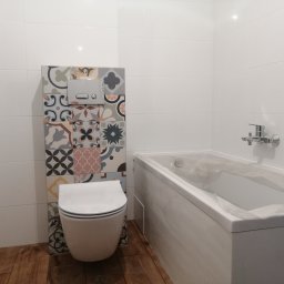 KTM-BUD JACEK LUBACZEWSKI - Biała łazienka z drewnopodobną podłogą, białe płytki na ścianach, zabudowa toalety z dekoracyjnymi płytkami, biała wanna zabezpieczona folią.